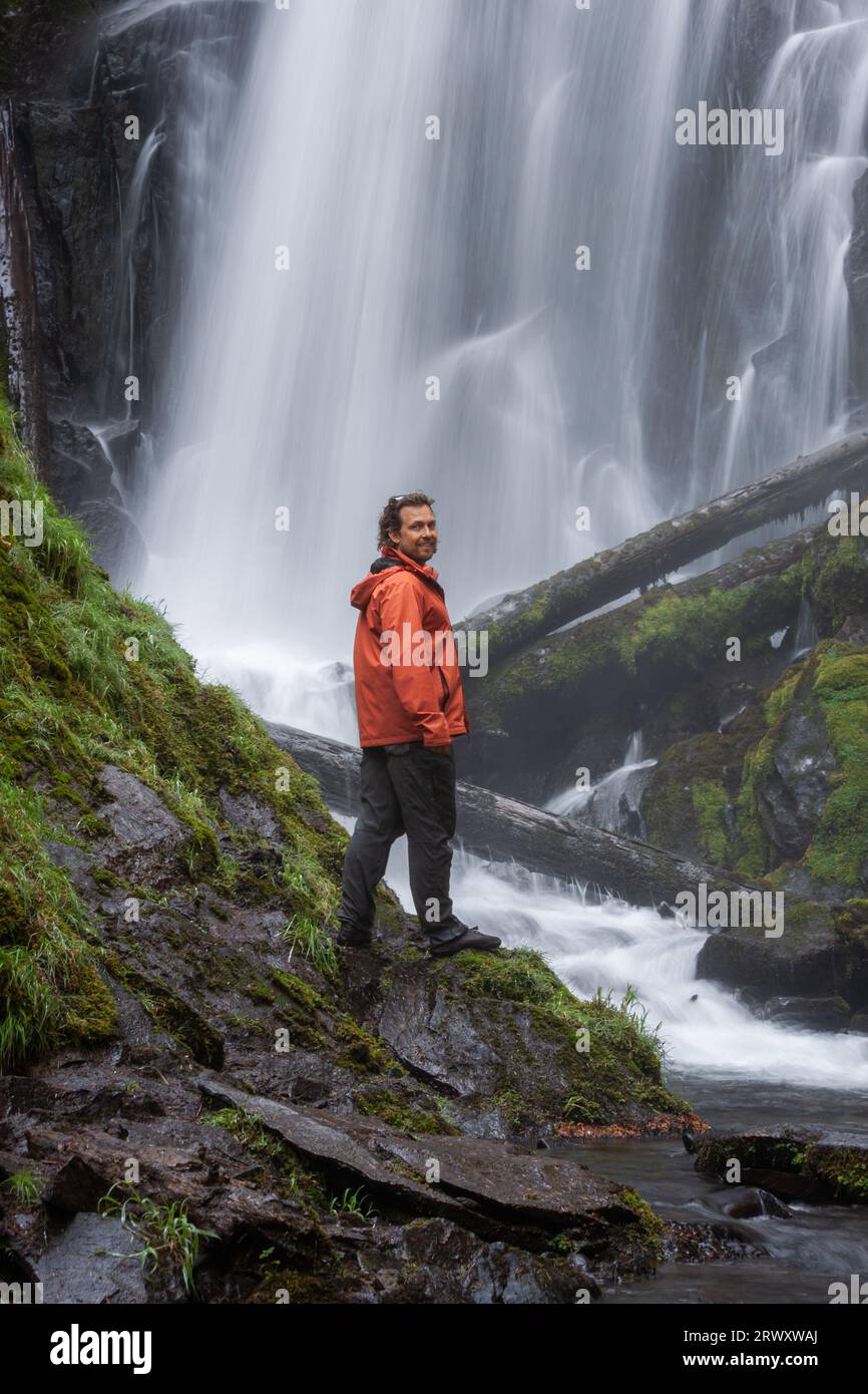 Wanderer tragen eine orangefarbene Jacke, die vor einem großen und wunderschönen Wasserfall im Wald von Oregon steht Stockfoto