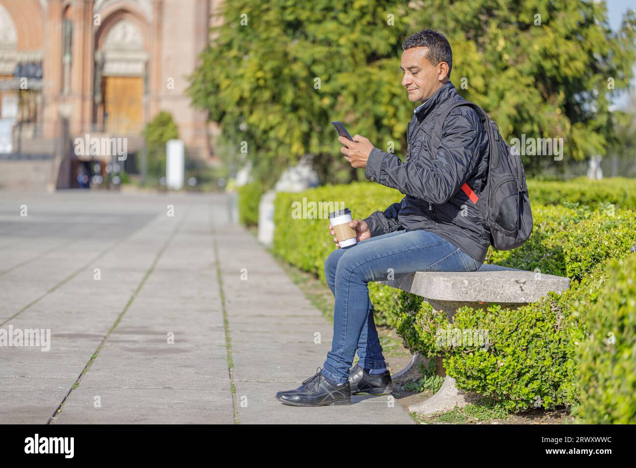 Junger Latino, der auf einer Bank in einem öffentlichen Park sitzt, sein Handy benutzt und einen Kaffee isst. Stockfoto