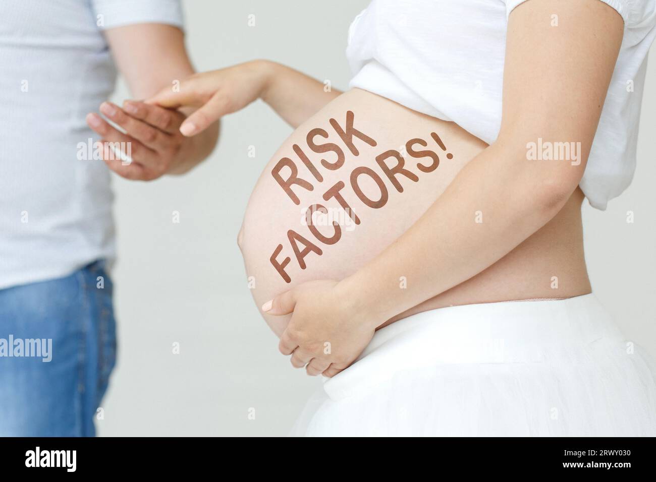 Eine schwangere Frau hält die Hand ihres Mannes auf dem Bauch mit der Inschrift - Risikofaktoren, ein Ausrufezeichen. Schwangerschaftskonzept. Stockfoto