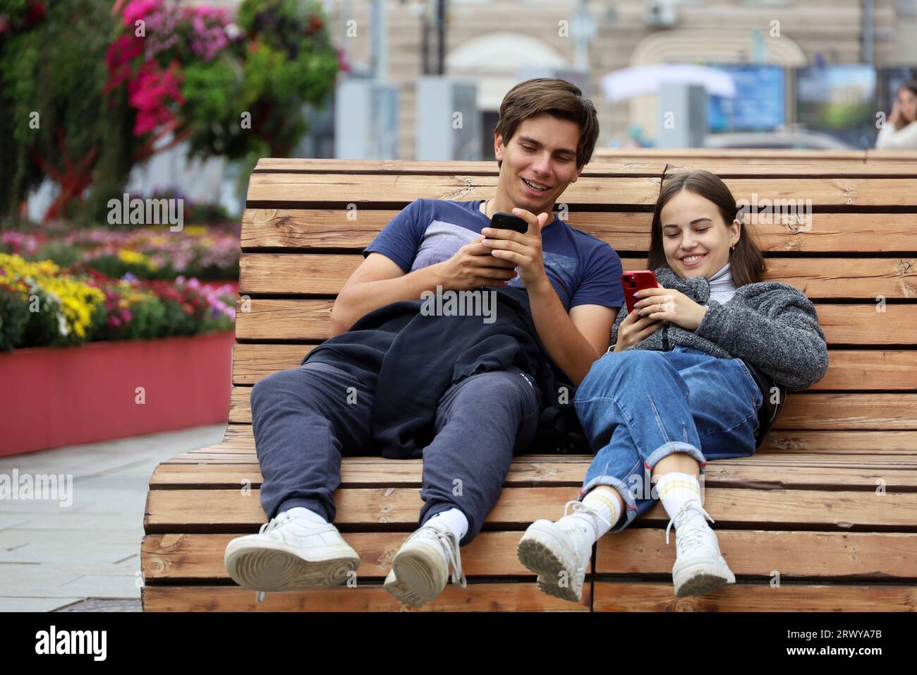 Glückliches junges Paar, das in der Herbststadt auf einem Smartphone-Bildschirm sitzt und auf einer Holzbank sitzt Stockfoto