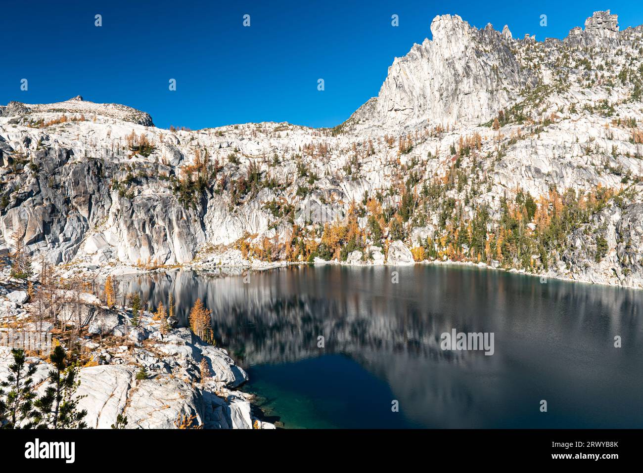 Granitklippen des Prusik Peak sind mit goldenen Lärchen übersät und umgeben das tiefe blaue Wasser des Viviane-Sees an einem knusprigen Herbsttag in The Enchantment Stockfoto