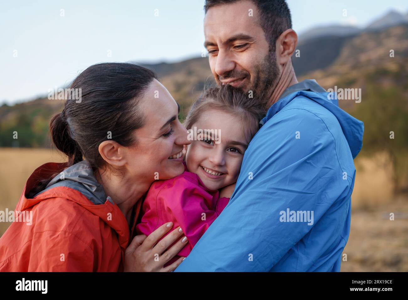 Mediterraner Familienmoment in den Bergen: Ein liebevoller Vater und eine liebevolle Mutter kuscheln ihre aufmerksame Tochter bei einem Outdoor-Abenteuer in den Bergen, während sie schaut Stockfoto