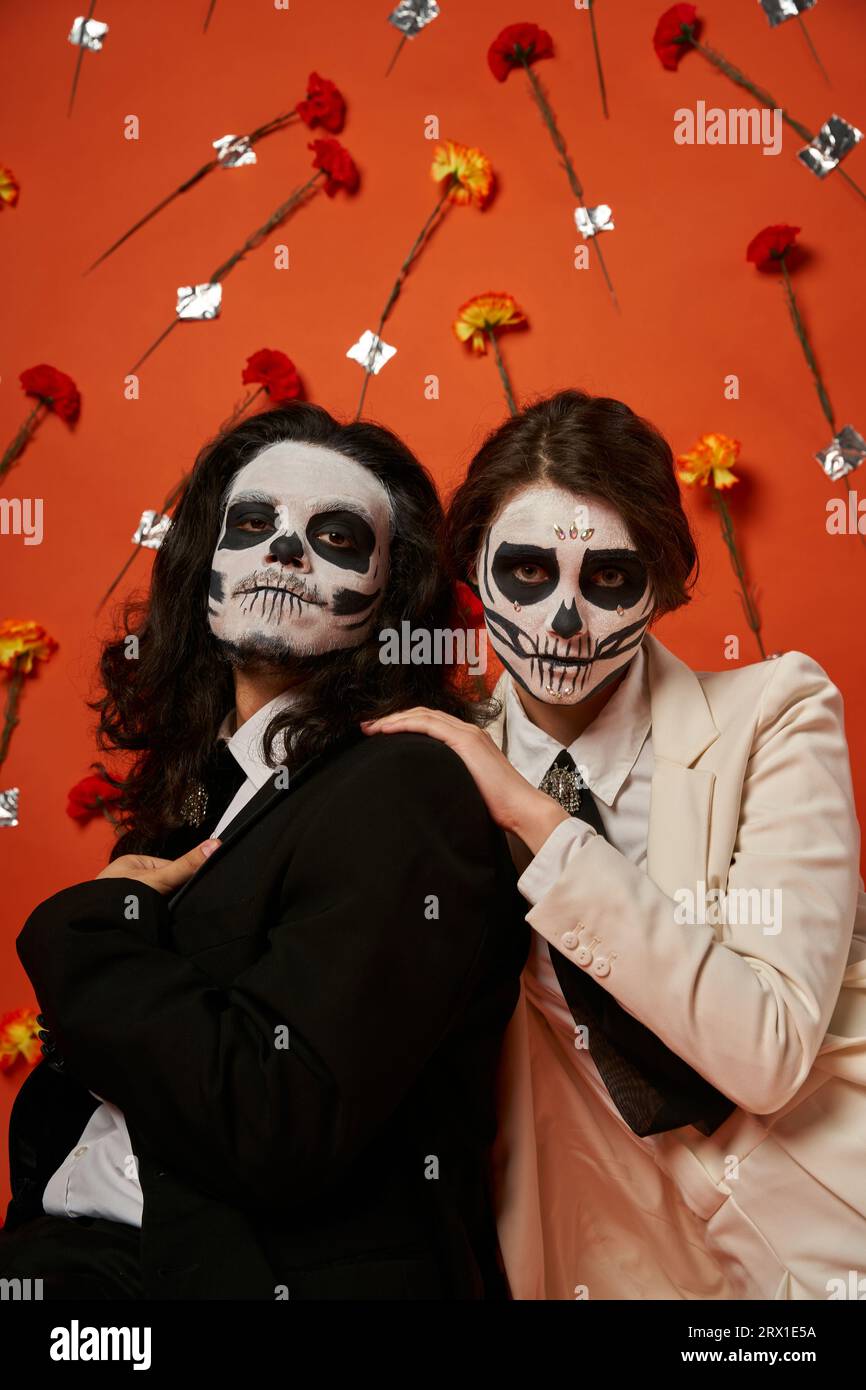 Gruseliges Paar in DIA de los muertos-Make-up und festliche Kleidung vor rotem Hintergrund mit Blumen Stockfoto