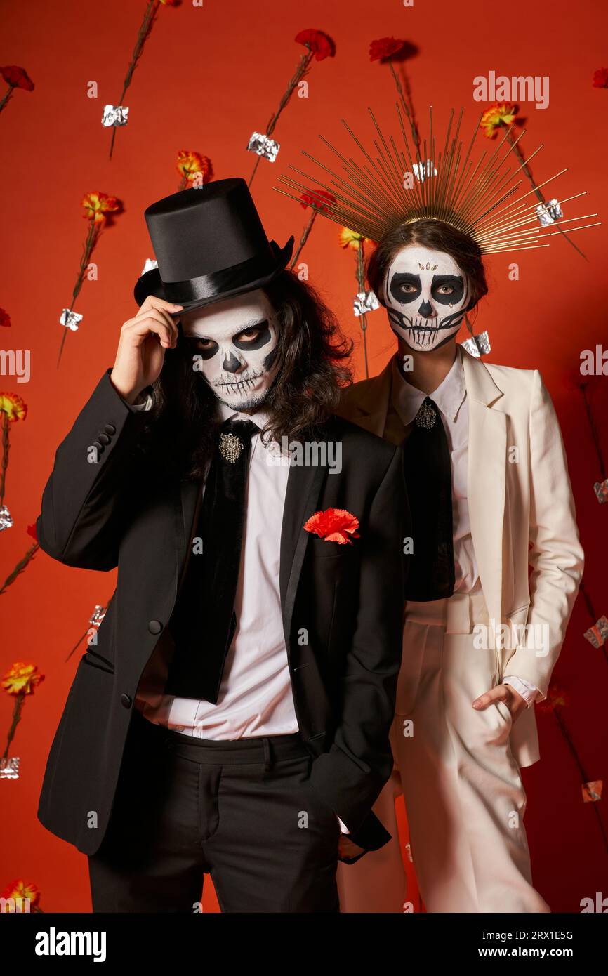 Elegantes Paar mit Totenkopf-Make-up und festlichem Outfit auf rotem Hintergrund mit Blumen, Day of Dead Party Stockfoto