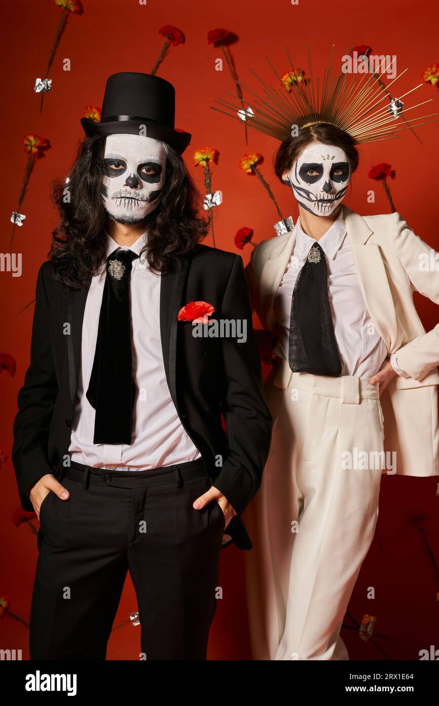 Ehepaar in DIA de los muertos-Make-up und festlichem Outfit mit roter Kulisse und Nelken Stockfoto