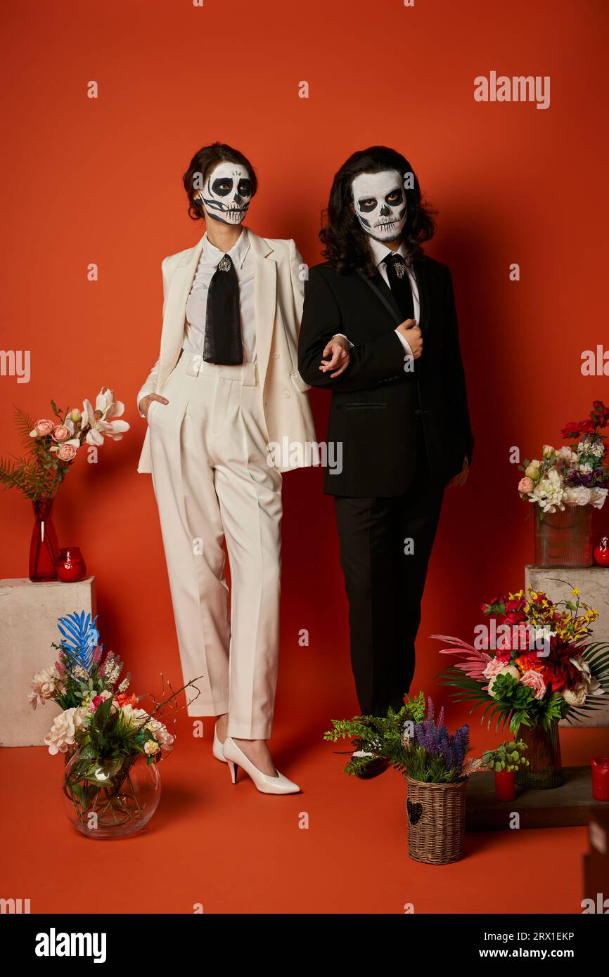 Paar in catrina Make-up und Anzüge in der Nähe von DIA de los muertos ofrenda mit Blumen auf rot Stockfoto