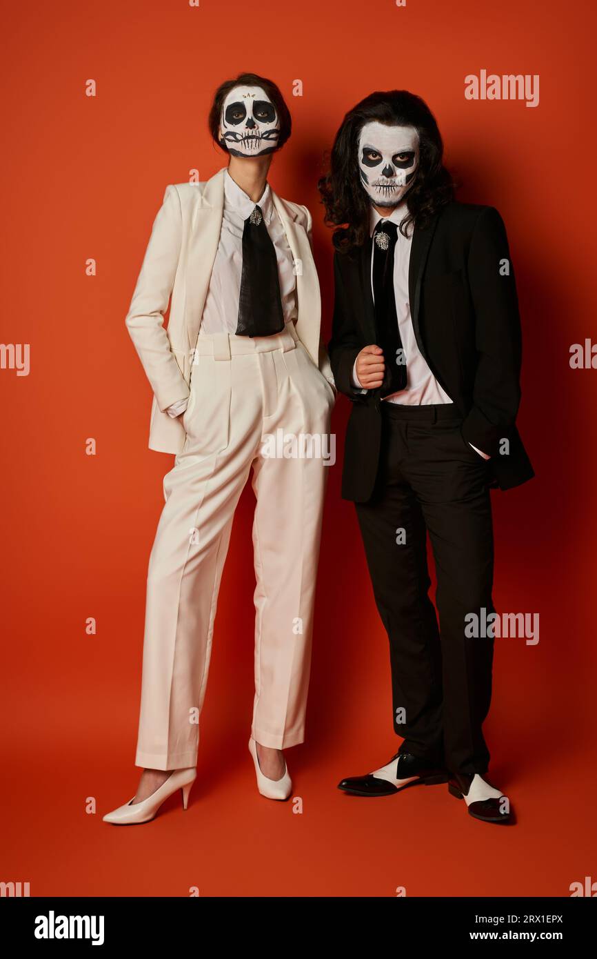 Ein stilvolles Paar in voller Länge mit Totenkopf-Make-up und eleganten Anzügen, die mit Händen in roten Taschen posieren Stockfoto