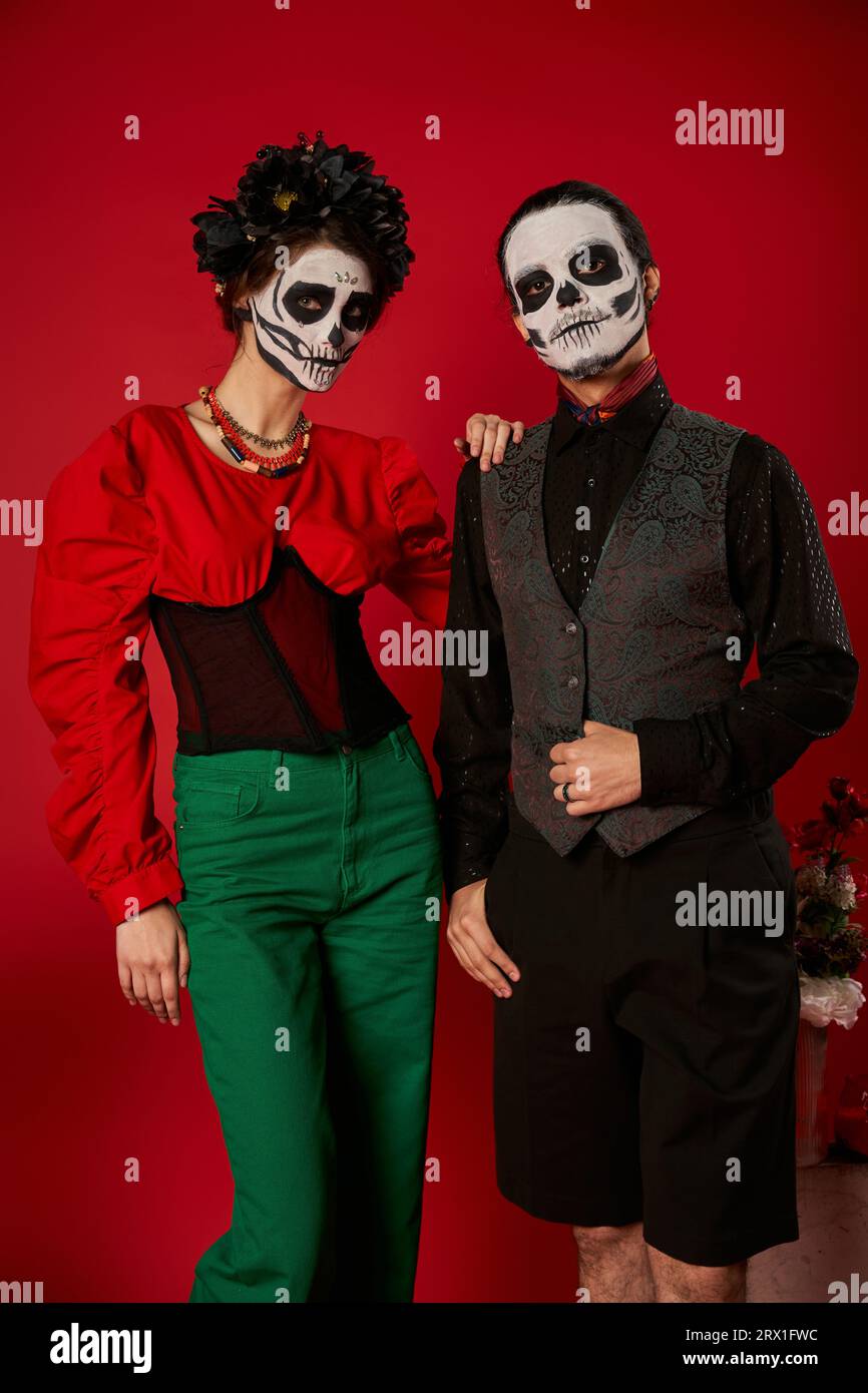 Elegantes Paar in DIA de los muertos-Make-up und festlicher Kleidung, das die Kamera in der Nähe von Blumen auf rot betrachtet Stockfoto