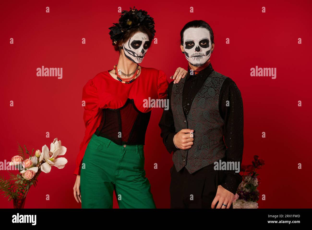 Das stylische DIA de los Muertos-Paar im Totenschädel-Make-up schaut auf die Kamera in der Nähe von Blumen auf rot Stockfoto
