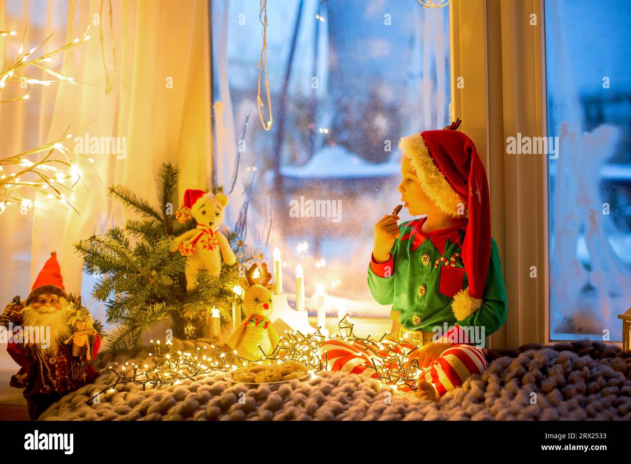 Niedliches Kleinkind in Pyjama, das auf einer wütsamen Decke neben einem Fenster sitzt und draußen auf ein Feuerwerk am Silvesterabend blickt Stockfoto