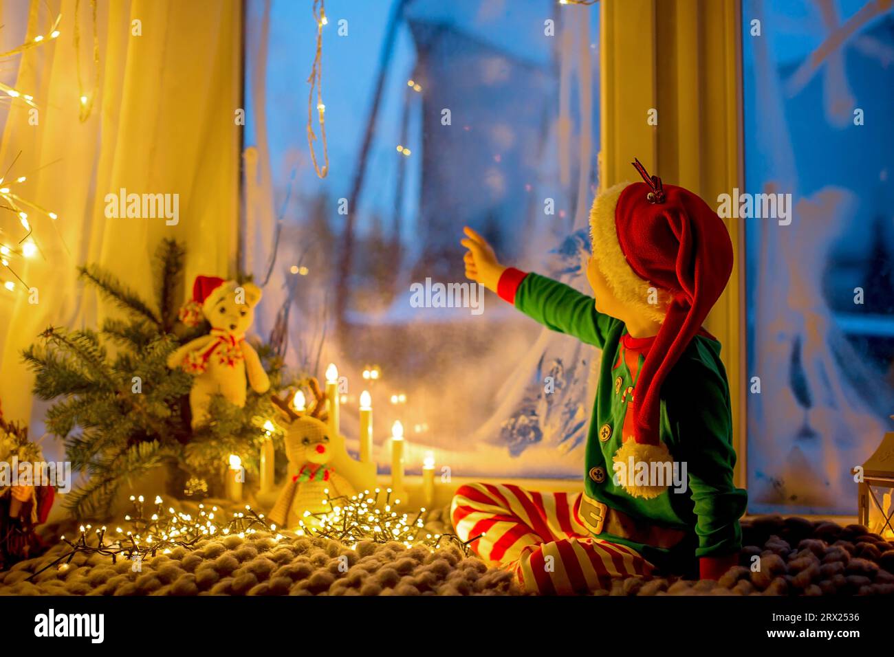 Niedliches Kleinkind in Pyjama, das auf einer wütsamen Decke neben einem Fenster sitzt und draußen auf ein Feuerwerk am Silvesterabend blickt Stockfoto