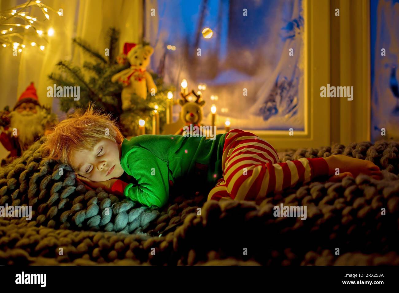 Niedliches Kleinkind im Pyjama, schlafend auf einer furiosen Decke neben einem Fenster, blaue Nacht draußen mit Feuerwerk am Silvesterabend Stockfoto