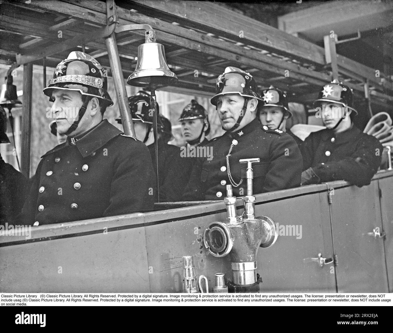 In den 1940er Jahren stürzen Feuerwehrleute der Kungsholmen-Feuerwehr in Stockholm in ihren offenen Feuerwehrauto. In Feueruniformen und Helmen mit Emblemen gekleidet, fahren sie aus der Feuerwache. Das Löschfahrzeug ist mit Leitern und einer Pumpenanlage ausgestattet. Eine Messingglocke ertönt während des Anrufs. Februar 1940. Kristoffersson, Ref. 57-3 Stockfoto