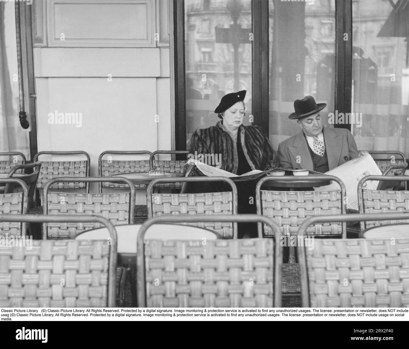 Paar in den 1950er Jahren Ein Paar, das auf einem Stuhl in einem Café im Freien sitzt, mit leeren Sitzen, bis auf die beiden. Die lesen die Tageszeitung. Sie trägt einen Pelzmantel und einen passenden Hut, er trägt einen Anzug mit Mantel und Hut. Schweden 1957 Stockfoto