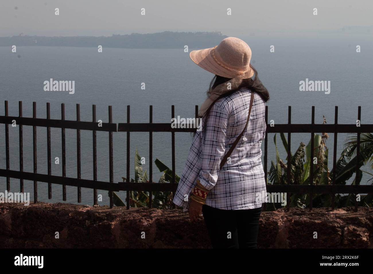 Blick von hinten auf die junge Frau, die das Meer von der umzäunten Terrasse aus bewundert. Stilvolle weibliche Reisende im Sommer Hut mit Blick auf das schöne Meer während des Urlaubs Stockfoto