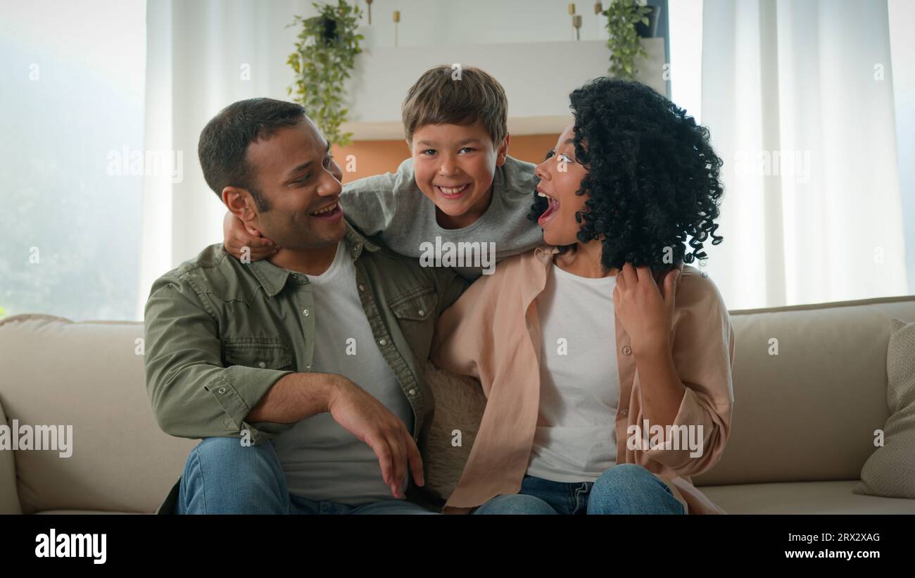 Liebevolle Eltern Erwachsene afroamerikanische Mutter und Vater umarmen Kuscheln auf Couch lustiger kleiner Junge adoptierter Sohn Kind Kind springt hinter Umarmung Mutter Stockfoto