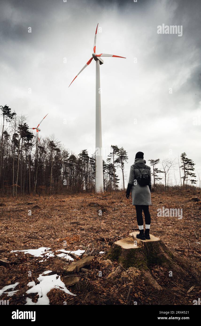 Frau steht und schaut auf Windkraftanlage in zerstörtem und entwaldetem Wald auf Baumstumpf bei schlechtem Wetter - Umweltproblem Stockfoto