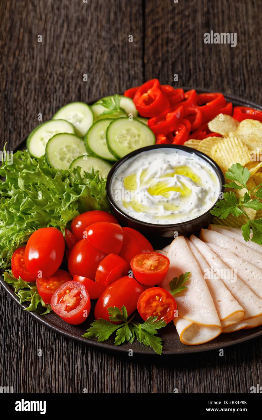 Nahaufnahme von geschnittenem Schinken, Kartoffelchips, Blattgemüse und Gemüse Snackplatte mit gesundem Joghurt Ranch Dip auf dunklem Holztisch, vertikale Ansicht Stockfoto