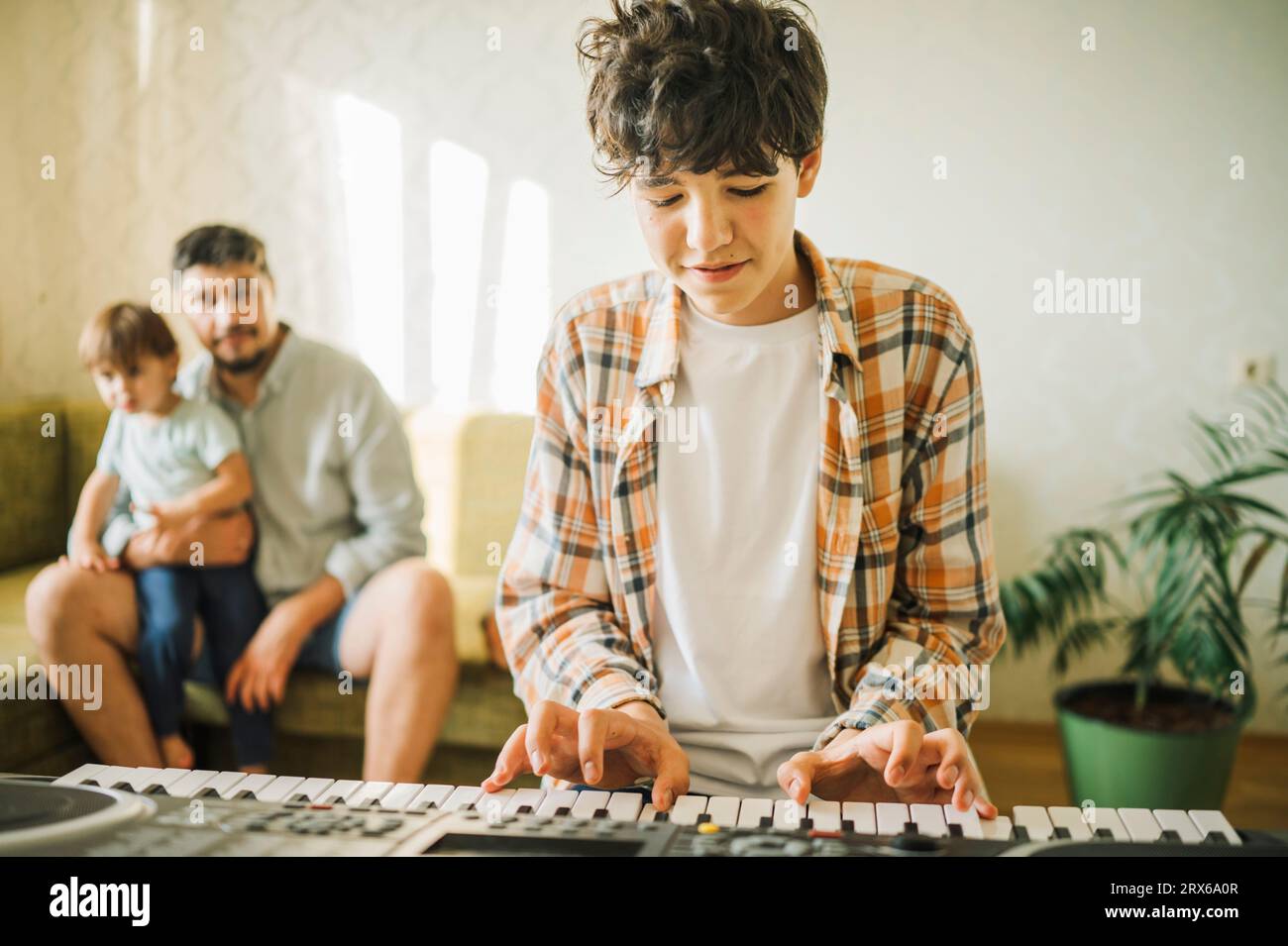Junge, der Klavier spielt, während Vater und Bruder im Hintergrund sitzen Stockfoto