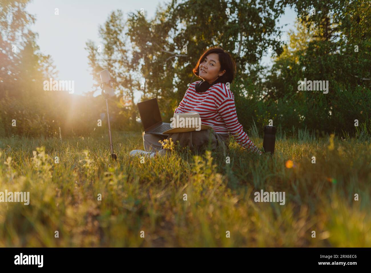 Lächelnde reife Frau mit Hut, der auf dem Feld auf Gras sitzt Stockfoto