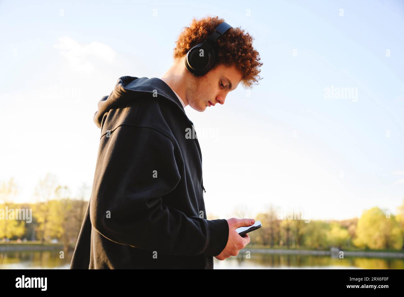 Teenager-Junge, der das Mobiltelefon nutzt und Musik über Kopfhörer im Park hört Stockfoto