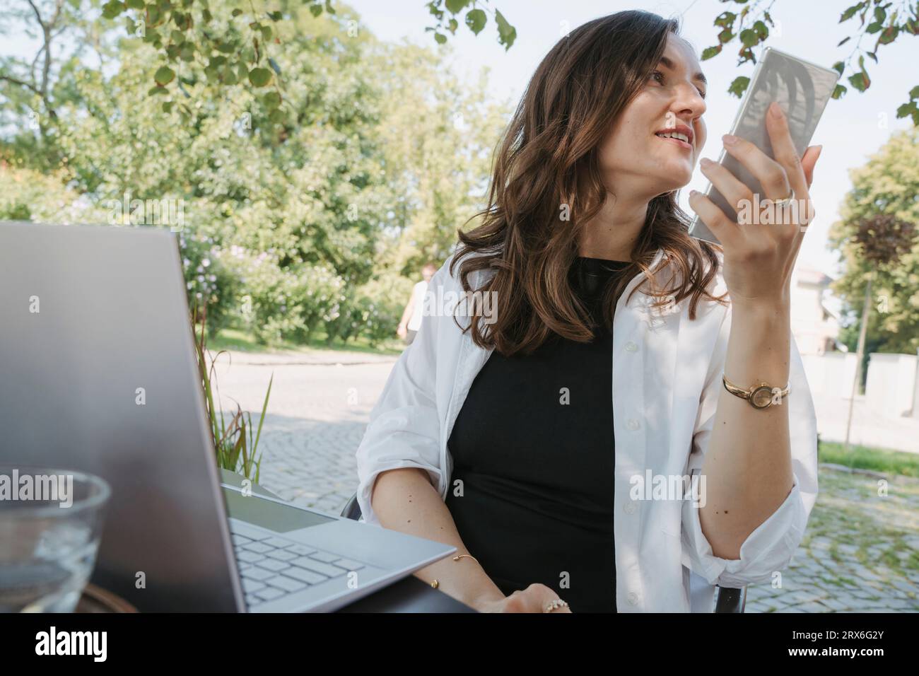 Lächelnde Geschäftsfrau, die Voicemail über das Smartphone am Tisch im Garten aufnimmt Stockfoto