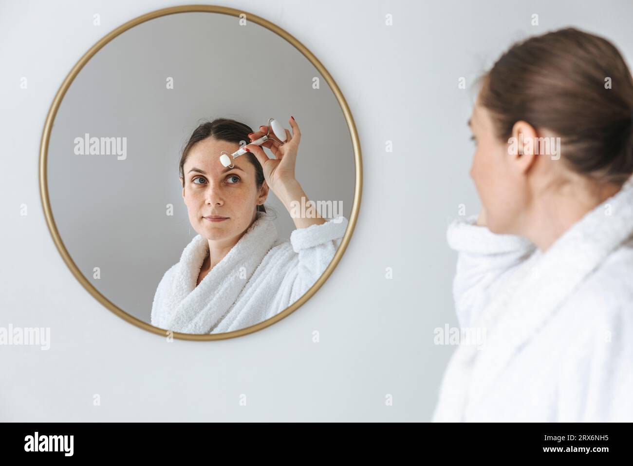 Lächelnde Frau mit Jadewalze im Gesicht und Blick in Spiegel zu Hause Stockfoto