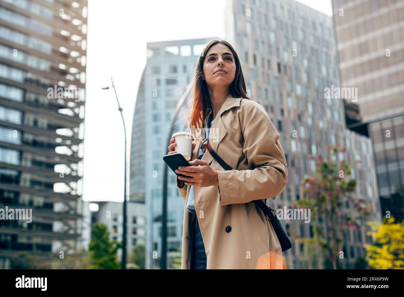 Nachdenkliche junge Frau, die mit Smartphone und Kaffeetasse steht Stockfoto