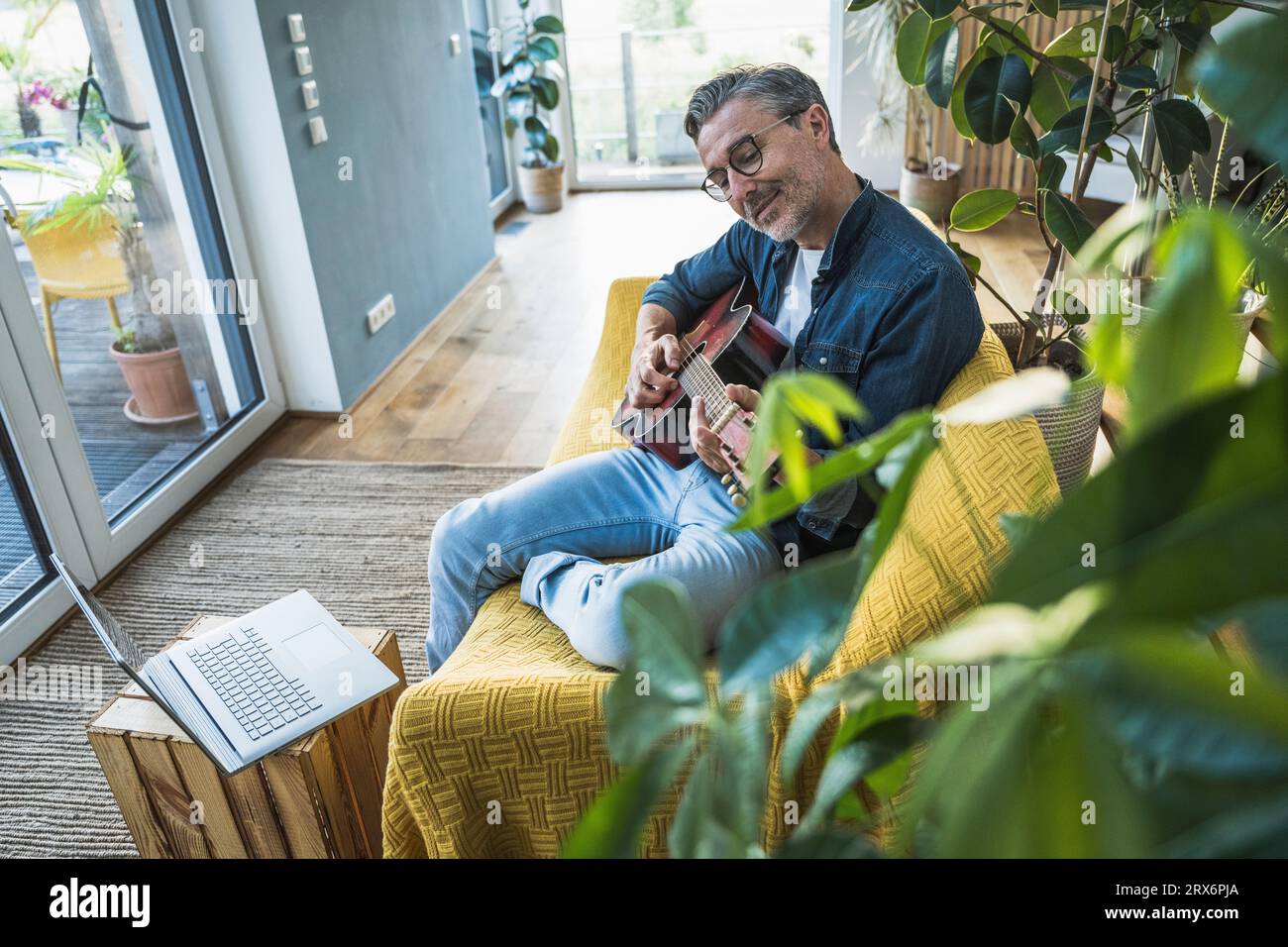 Mann, der zu Hause Gitarre spielt, sitzt auf dem Sofa am Laptop Stockfoto
