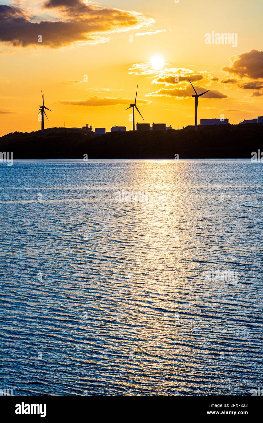 Der Sonnenuntergang hinter Windturbinen in der ehemaligen Ölraffinerie Gulf in Waterston, Milford Haven in Pembrokeshire, West Wales, Vereinigtes Königreich Stockfoto