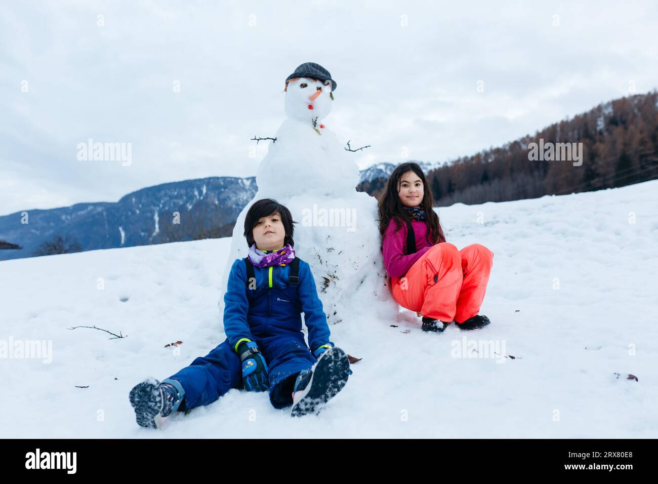 Zwei Kinder sitzen neben dem Schneemann, den sie auf dem schneebedeckten Berg gemacht haben Stockfoto