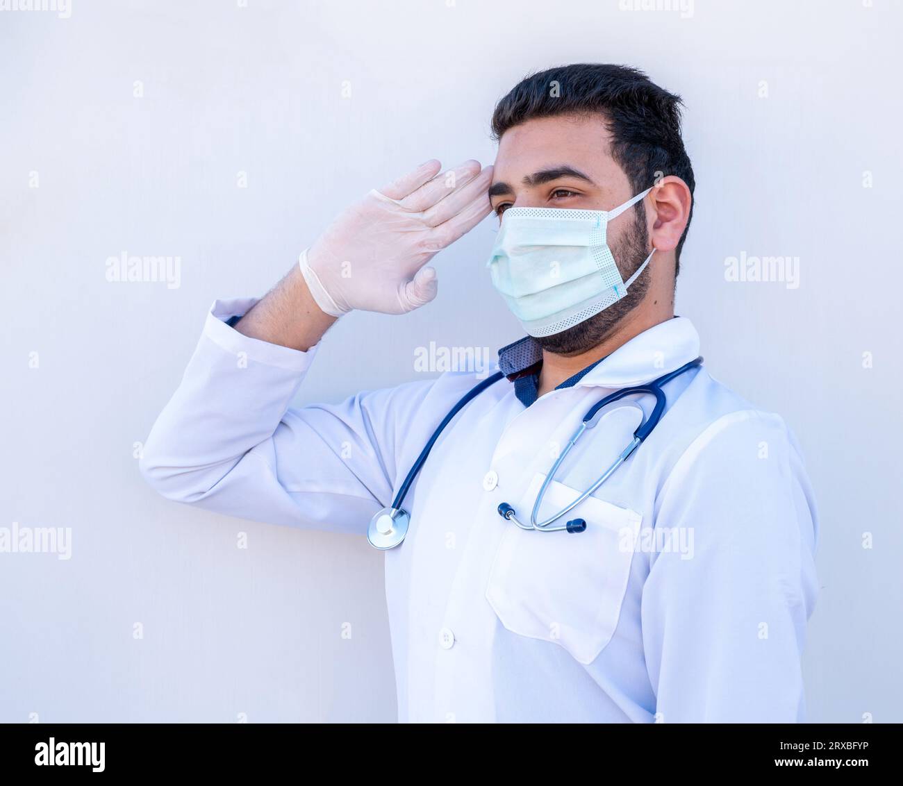 Doktor, der militärischen Gruß auf isoliertem weißem Hintergrund tut und sein Stethoskop während des Gesundheitstages trägt Maske Stockfoto