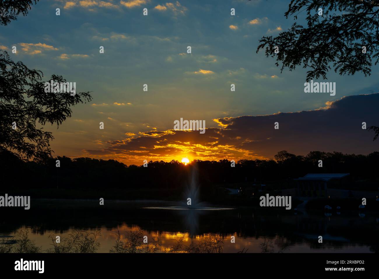 Ein Sonnenaufgang im September spiegelt sich auf Wolken und einem Teich mit Springbrunnen. Stockfoto