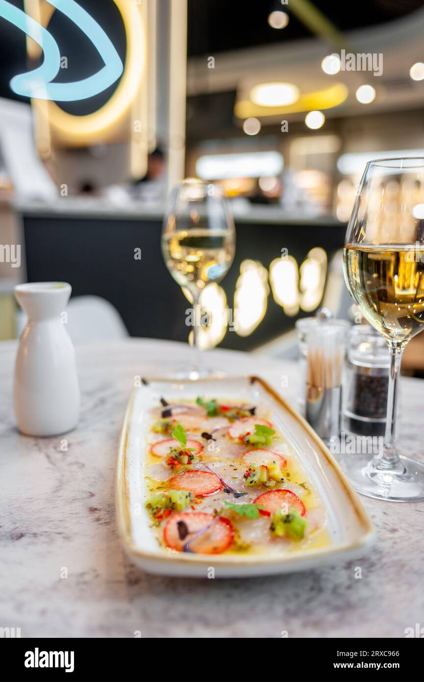 Wolfsbarsch-Ceviche mit Erdbeeren, Kiwi und Mangosalsa auf einem Marmortisch in einem Restaurant und einem Glas Weißwein Stockfoto