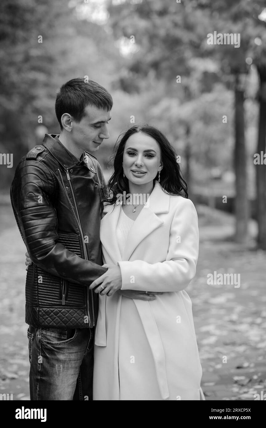 Ein Mann umarmt seine schwangere Frau. Ein Mann in einer schwarzen Jacke umarmt und küsst eine Frau in einem weißen Mantel. Herbst Stockfoto