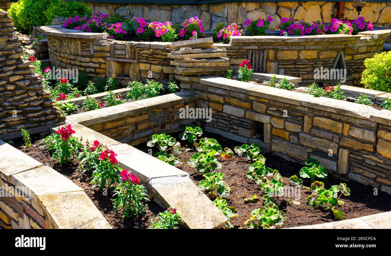 Landschaftsgestaltung mit Stützmauern und Blumenbeeten im Hinterhof des Hauses, Landschaftsgestaltung eines gehobenen Hausgartens mit Steinmauern im Sommer. Blumen und pla Stockfoto