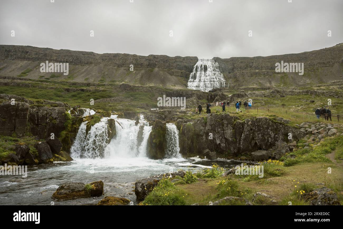 Winzige Touristen, die den Weg zum Boden des Dyndanji-Wasserfalls in Westfjords Island mit Baejarfoss-Wasserfall im Vordergrund und A erklimmen Stockfoto