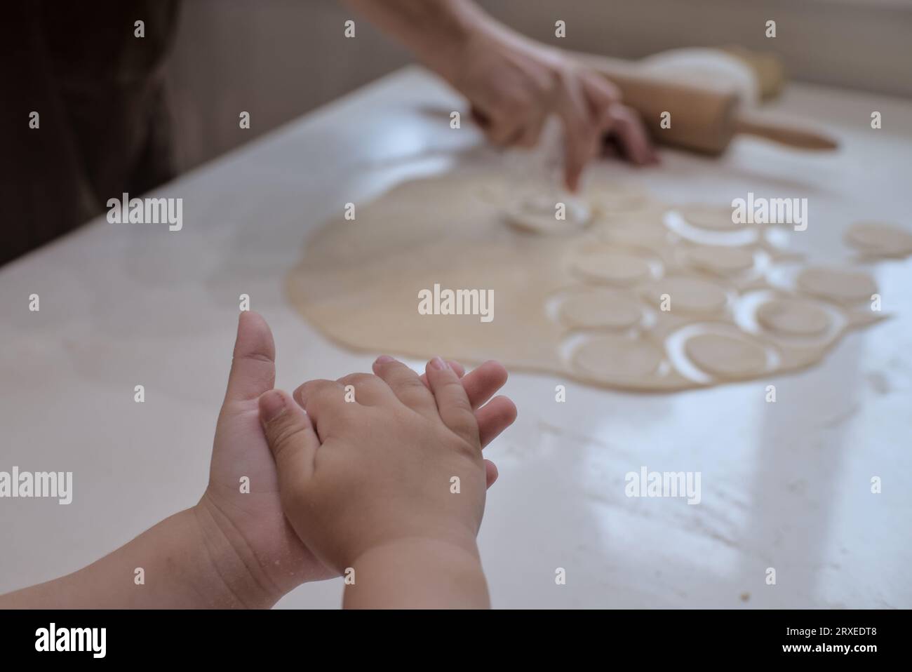 Kleine Kinderhände halten Teig, während die Erwachsene Frau im Hintergrund Essen zubereitet. Stockfoto