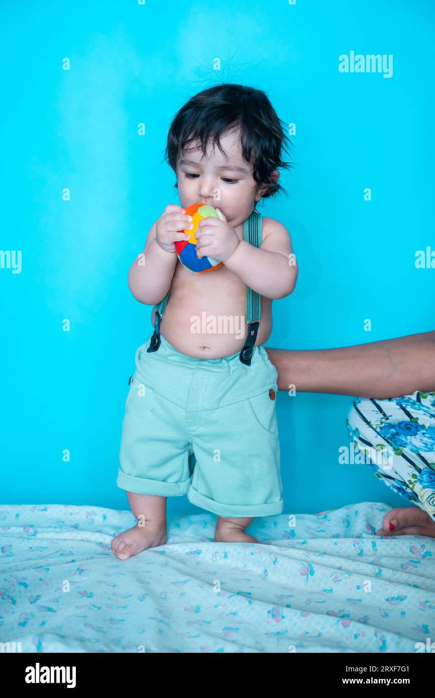 Lächelnder sechs Monate alter süßer indischer Junge, der versucht, während Mutter isoliert über blauem Studiohintergrund hält, glückliches Kind mit schwarzem Haar. Stockfoto
