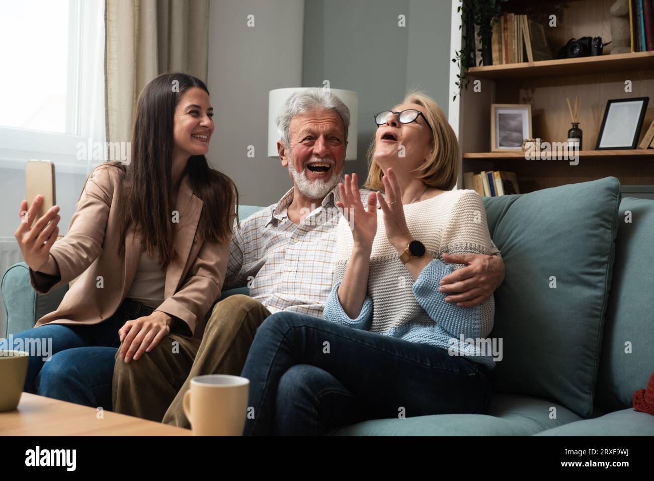 Die Tochter oder Enkelin, die ihre Eltern besucht, ein älteres Paar oder Großeltern macht ein Foto von ihnen mit ihrem Handy und nimmt ein Selfie als Erinnerung auf Stockfoto