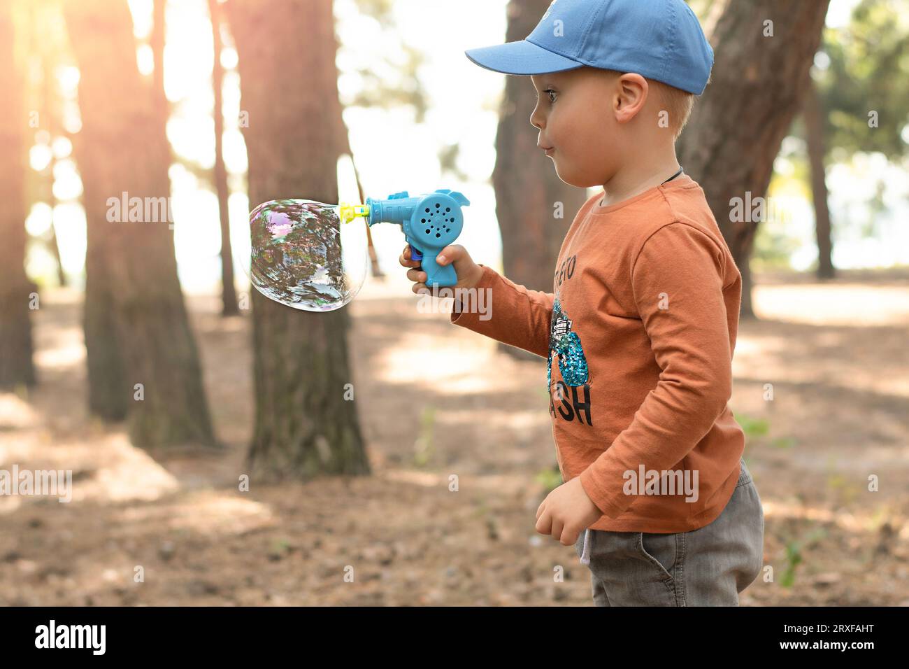 Kinder. Ein kleiner gutaussehender und emotionaler Junge in blauer Kappe und roter Jacke spielt mit Seifenblasen in der frischen Luft im Wald. Stockfoto