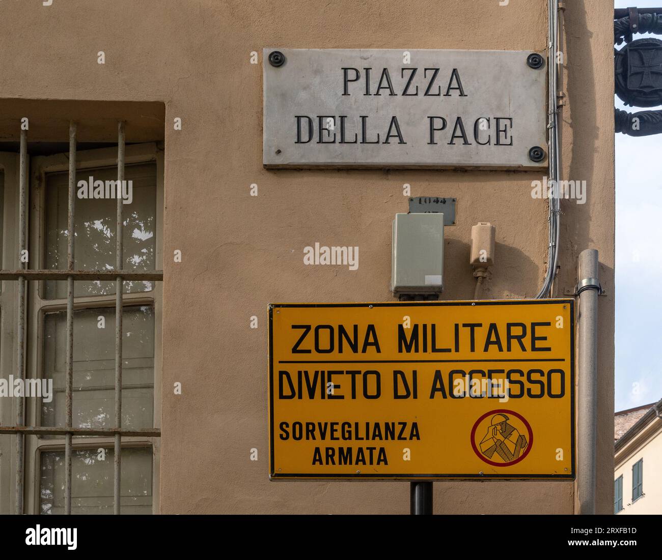 Kontrast zwischen dem Zeichen der Piazza della Pace („Friedensplatz“) und einem Schild mit der Aufschrift „Militärzone, kein Zugang, bewaffnete Überwachung“, Parma, Italien Stockfoto