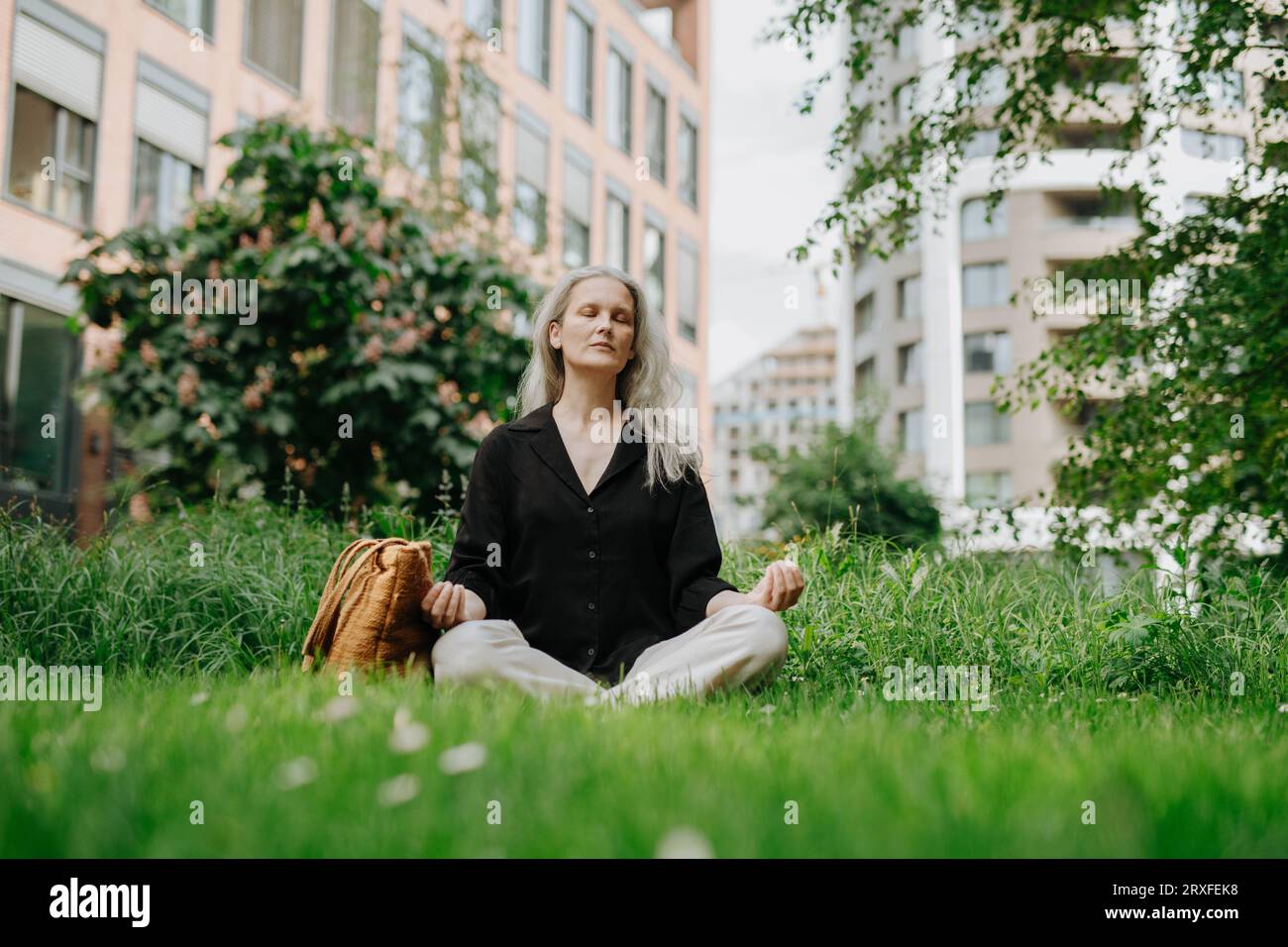 Porträt einer schönen lächelnden Frau mit grauem Haar, das auf dem Gras im Stadtpark meditiert. Stockfoto
