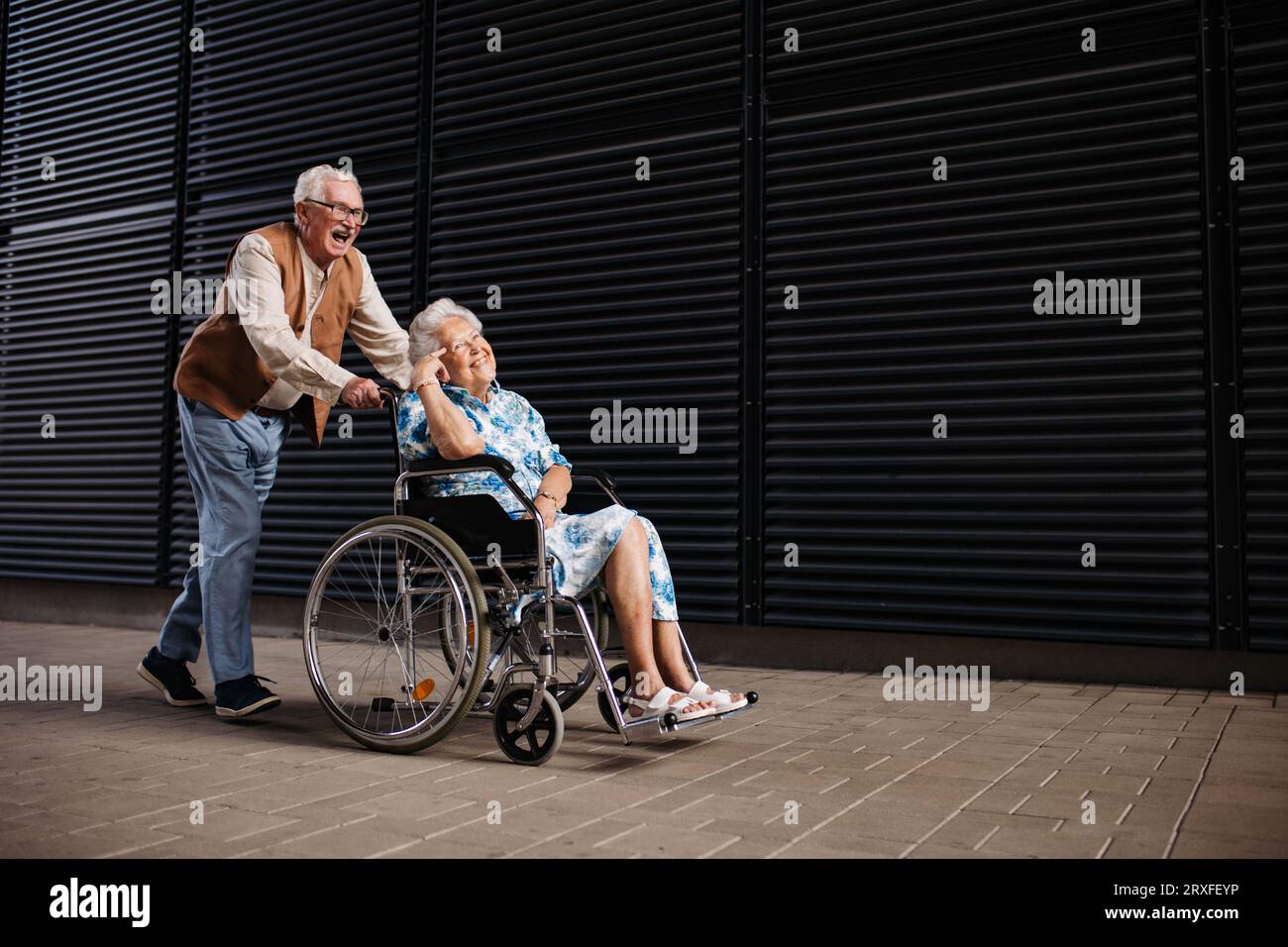 Älterer Mann, der seine lächelnde Frau im Rollstuhl schiebt. Stockfoto