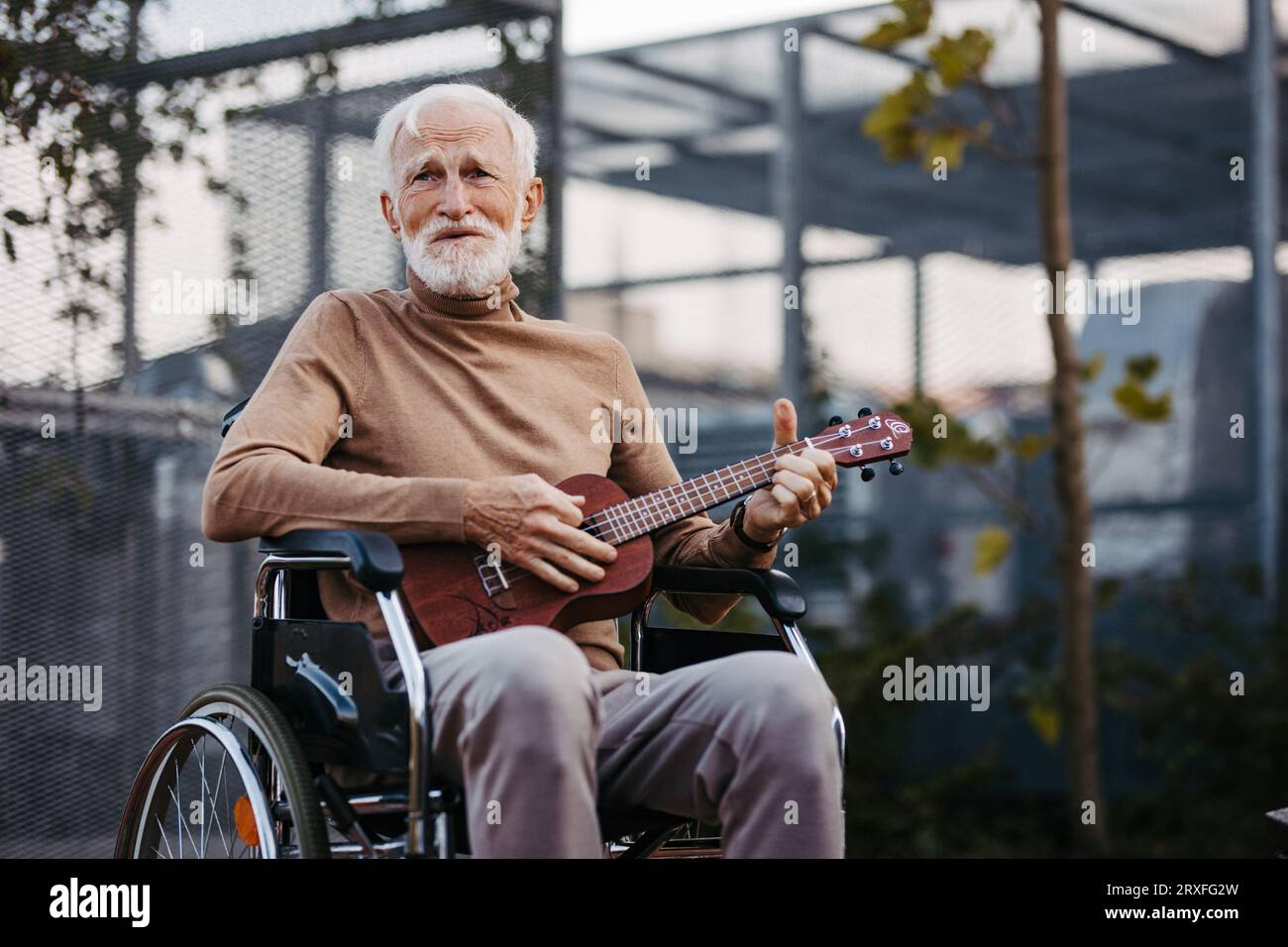 Älterer Mann, der draußen im Rollstuhl sitzt und die Ukulele spielt. Stockfoto