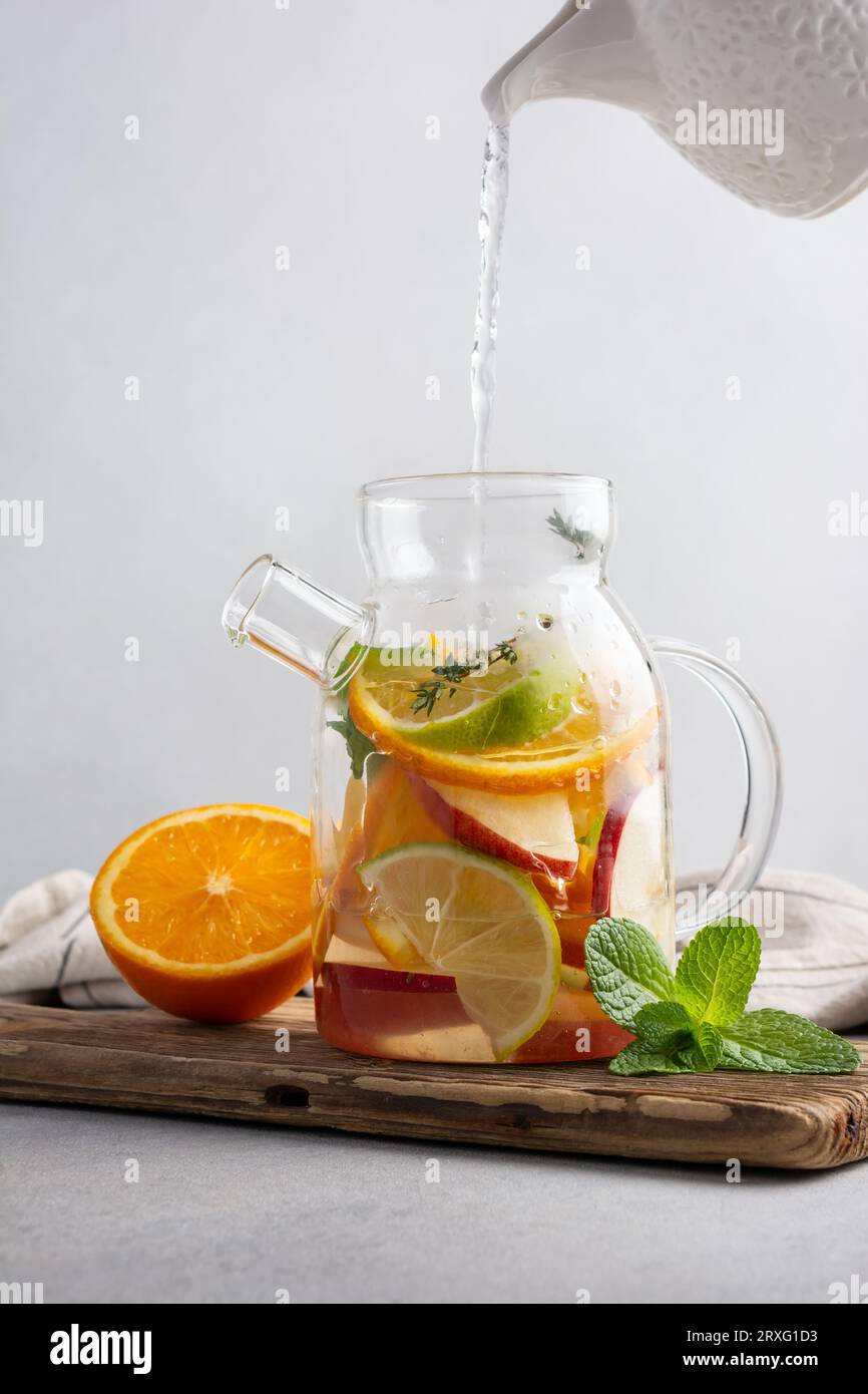 Hausgemachter, gesunder heißer Obsttee mit frischen, reifen Orangen, Apfeln, Minzblättern und Thymianzweigen in Glas-Teekanne oder Wasserkocher auf grauem Küchenhintergrund Stockfoto