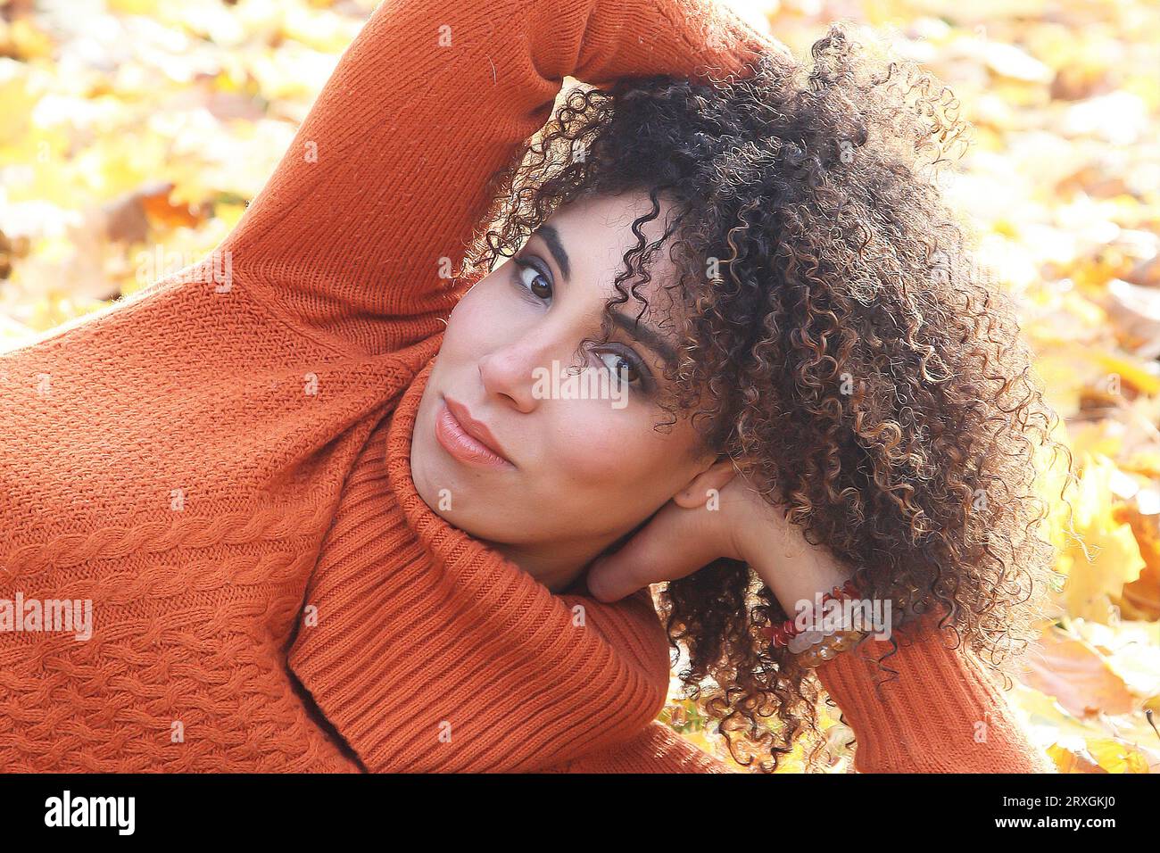 Lockige Frau mit orangefarbenem Pullover posiert mit den Herbstblättern im Hintergrund Stockfoto