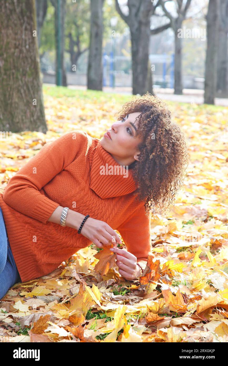 Lockige Frau mit orangefarbenem Pullover posiert mit den Herbstblättern im Hintergrund Stockfoto