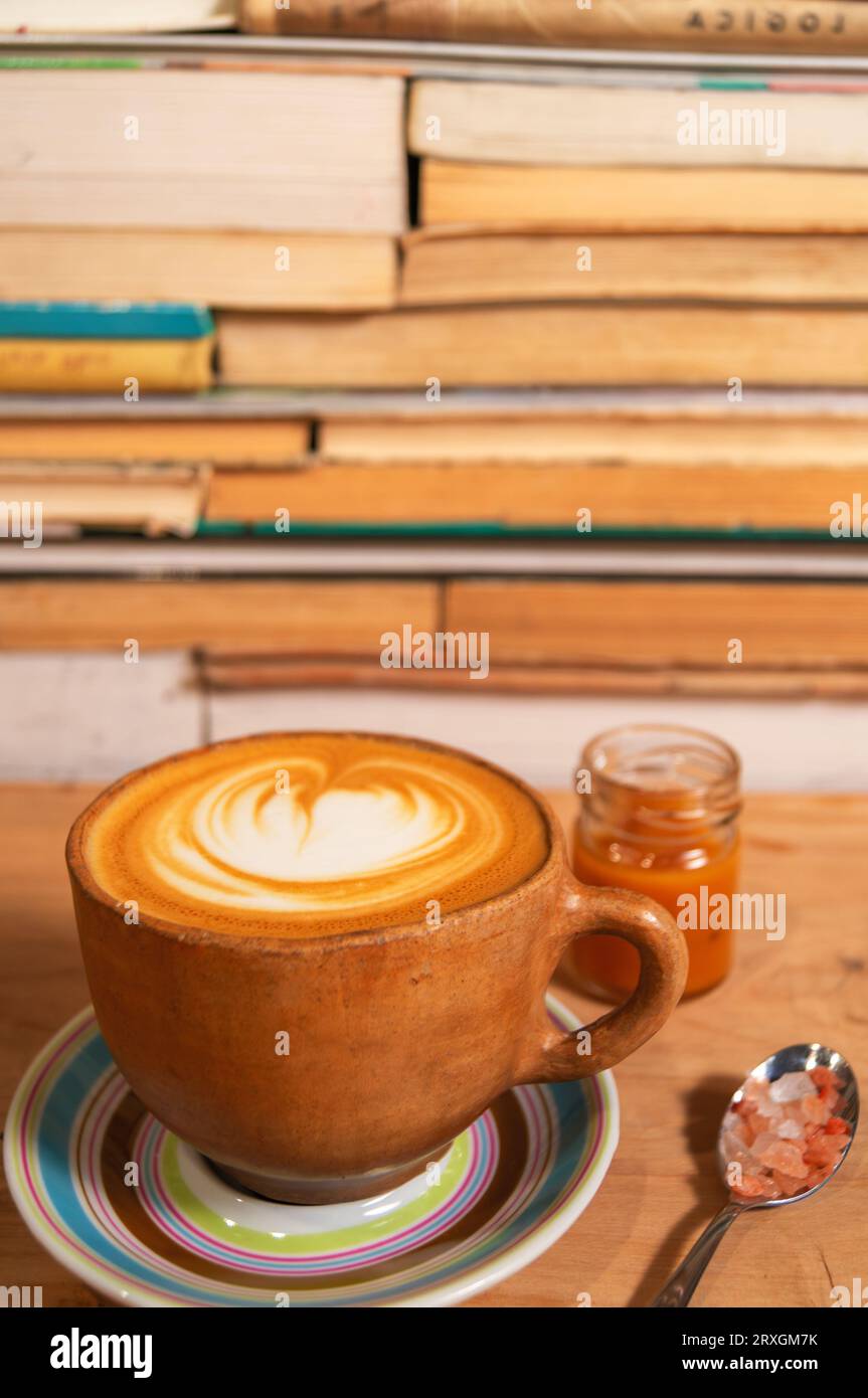 Rustikaler ockerfarbener Cappuccino-Becher mit Karamell und Himalaya-Salz, geformter Schaum auf Teller, Holz und Buchhintergrund - Taza de Capuchino rústica ocre con caram Stockfoto