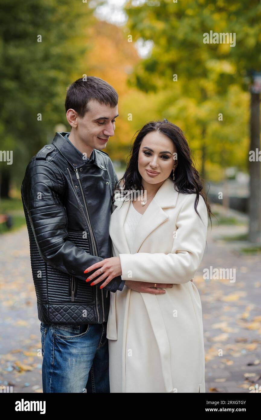 Ein Mann umarmt seine schwangere Frau. Ein Mann in einer schwarzen Jacke umarmt und küsst eine Frau in einem weißen Mantel. Herbst Stockfoto